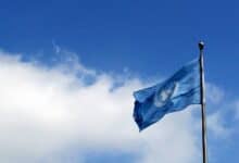 The image of United Nations flag waving against a bright blue sky with scattered clouds.