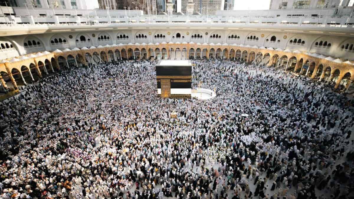 A large crowd of pilgrims gathers around the Kaaba inside the Masjid al-Haram in Makkah during Umrah. The mosque’s white marble flooring and arched colonnades surround the central black-draped structure as worshippers perform tawaf.