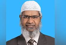 Zakir Naik pictured in a suit and white cap against a blue background.