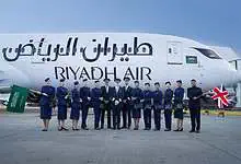 Riyadh Air flight crew and pilots pose in front of a Boeing 787 aircraft at Heathrow Airport, holding Saudi and British flags.