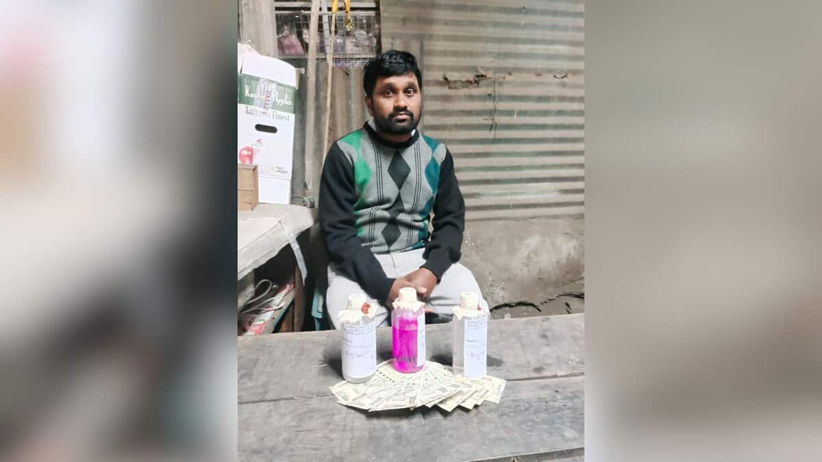 A man wearing a dark sweater with green and gray argyle pattern sits at a table with three bottles containing liquids (two clear, one pink) and Indian rupee notes arranged in front of him. The setting appears to be a small shop or workspace with corrugated metal walls and shelving visible in the background
