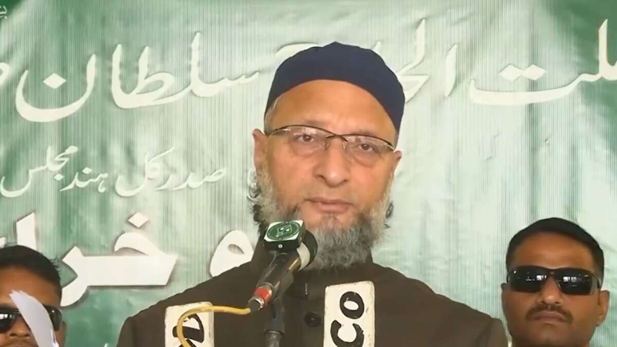 Man wearing black cap and glasses with a beard speaks at microphones during a press conference, flanked by two men in sunglasses. Green banner with Arabic and Urdu text visible in background.