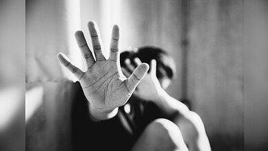 A person holds up their hand toward the camera in a defensive "stop" gesture while their face is obscured in the blurred background, conveying distress or a plea for help.