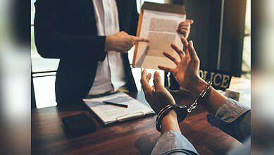 A person in handcuffs sits at a table during what appears to be a legal consultation, while someone in professional attire stands nearby holding a document. Law books and papers are visible on the wooden table surface.