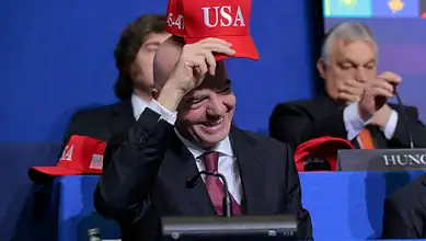 American delegate smiling and adjusting his red USA cap during a formal event.