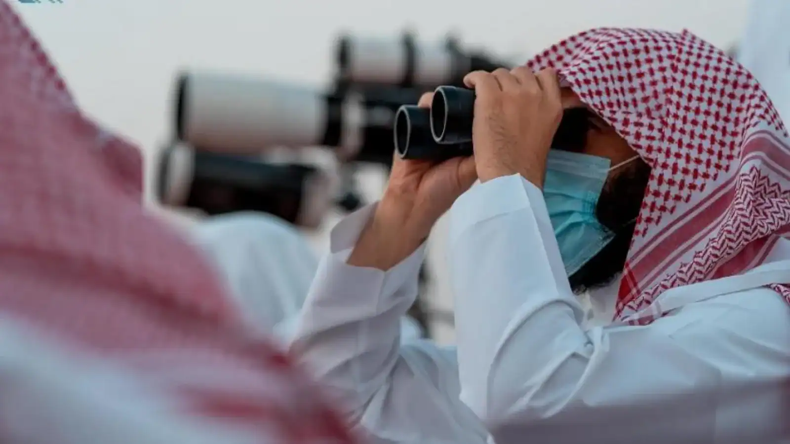 Saudi moon sighting committee member uses binoculars to observe the sky while wearing traditional attire and a face mask.