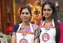 Two women chefs wearing aprons with MasterChef logo in a warm, decorated kitchen setting.