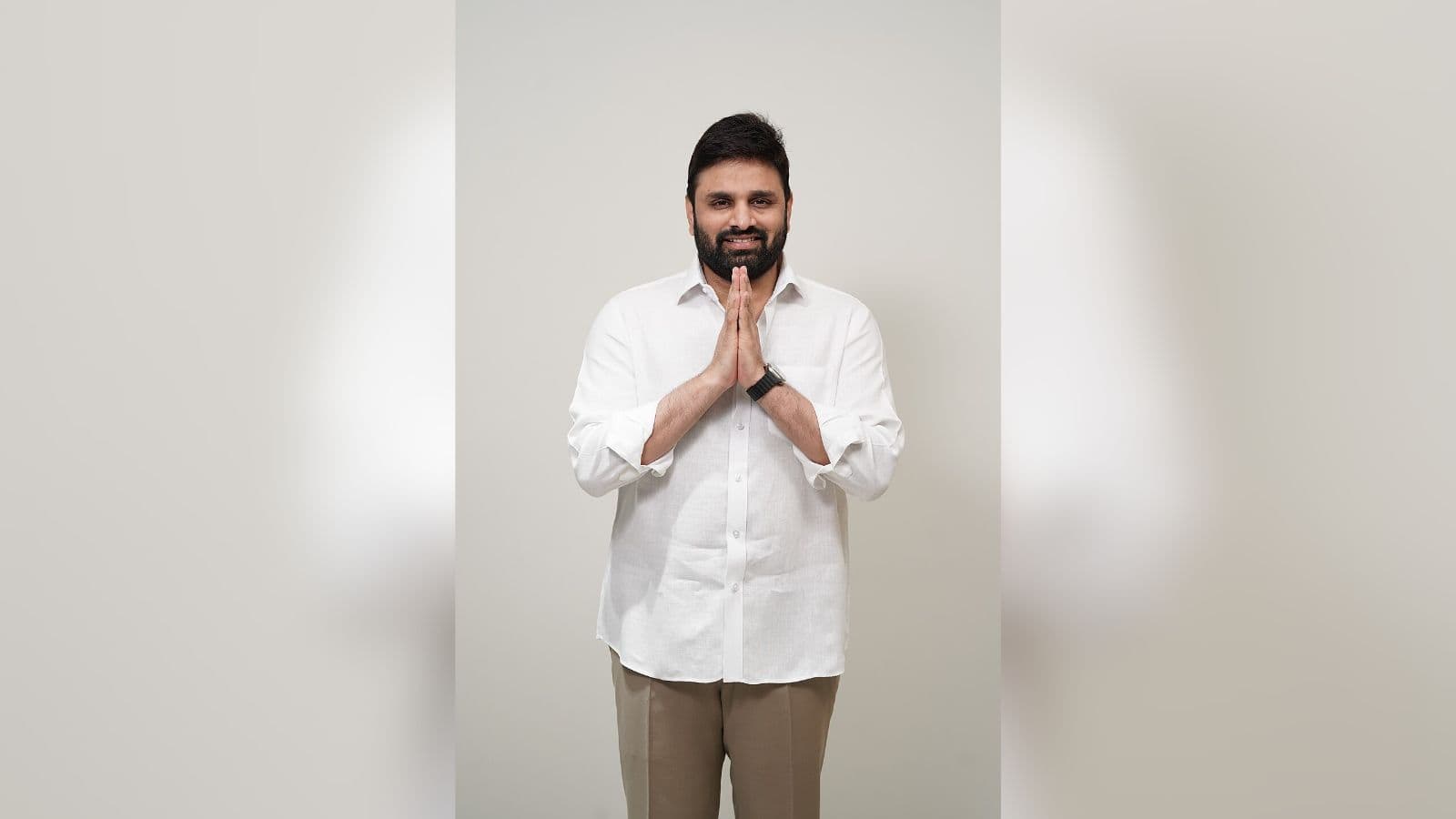 An image of a smiling man with hands folded in a gesture of greeting or respect, standing against a plain.