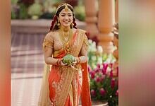 Rashmika Mandana dressed in vibrant traditional Indian attire, smiling and holding a decorative item duri.