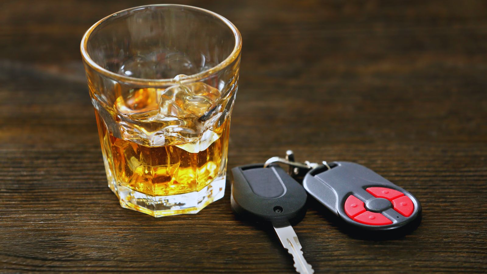 Image shows A glass of whiskey on the rocks beside car keys on a wooden surface.