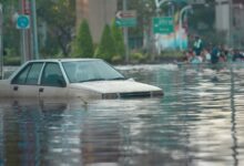 Image shows A white car nearly submerged in floodwater on a city street as onlookers watch from higher ground nearby.