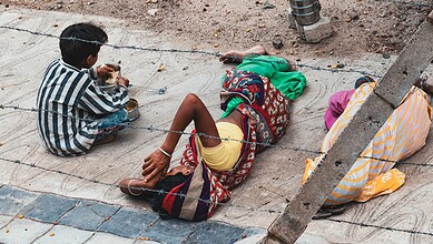 Child begging on the street, sitting near a woman lying on the ground, highlighting social issues and cas.