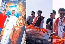 A man in traditional attire addresses a crowd, holding a framed picture, during a public event at Siasat.
