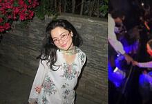 Hania Aamir smiling at a social gathering, wearing glasses and a floral dress, with a lively background.