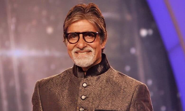 Amitabh Bachchan smiling in a formal traditional outfit, wearing glasses, on a stage with a blurred backg.