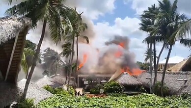 Resort on fire with thick smoke and flames, surrounded by palm trees in a tropical setting.