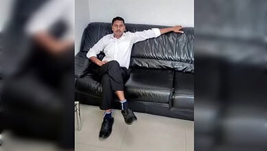 Man sitting on a black leather sofa, wearing a white shirt and black pants, in an indoor setting.