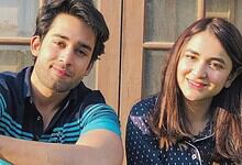 Young man and woman smiling outdoors in front of a wooden window.