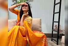 Rashmika Mandanna wearing a yellow saree, sitting on a sofa, smiling and shielding her eyes from the sun.