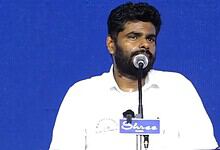 Annamalai speaking at a public event with a blue background and microphone.