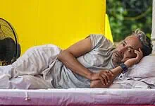 Man lying on bed in a yellow tent, appearing in pain and resting his head on his hand.