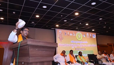 Speaker at Khairatabad Ganesh event holding a white bag, with a large screen behind him.