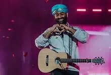 Arijit Singh performing on stage with a guitar and a blue turban, smiling and making a heart shape with.