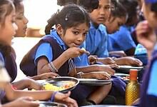 Young schoolgirls in blue uniforms eating a midday meal together, seated in a row with steel plates of rice.