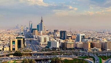 A panoramic view of Riyadh's skyline with modern skyscrapers and highways under a partly cloudy sky.