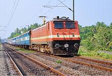 Indian Railways electric locomotive pulling passenger coaches through lush green countryside under overhead power lines.
