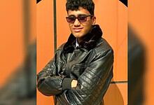 Young man wearing sunglasses and a leather jacket with arms crossed, smiling confidently.
