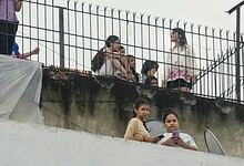 Group of young women and girls on a rooftop, some using mobile phones, with a metal fence in the scene.