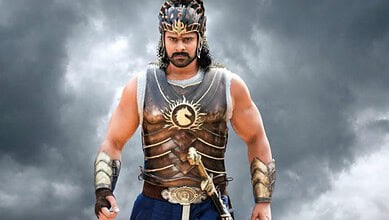 Prabhas as Baahubali wearing armor and crown, standing confidently against stormy sky.