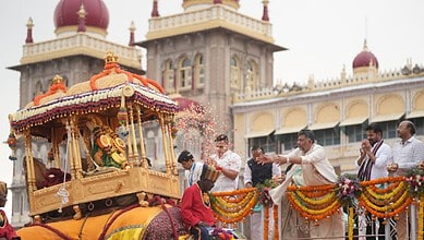 People performing a ritual during Mysuru Dasara with a decorated chariot in the background.