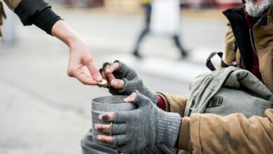 A midsection of woman giving money to homeless beggar man sitting in city.