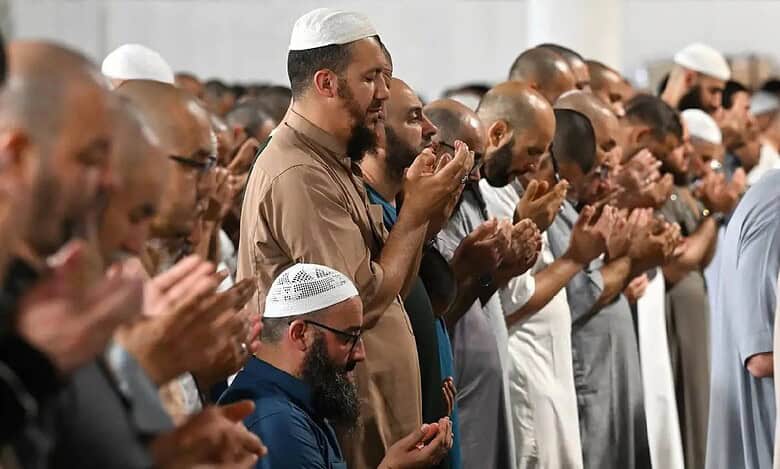 This image captures a group of Muslim worshippers standing in prayer at Makkah’s Grand Mosque during Isha and Taraweeh on the 29th night of Ramzan 1446 AH. The men, dressed in traditional attire such as thobes and kufis, are standing in rows with their hands raised in dua.