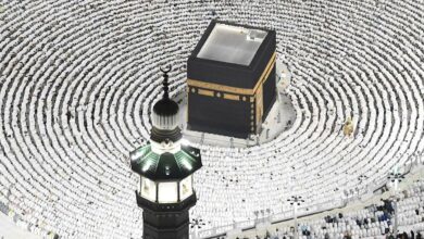 A crowd in white surrounds the Kaaba, a black cubic structure with gold details, in a lit courtyard at night. A green-lit minaret is in the foreground, during a religious gathering, likely Haj or Umrah in Mecca, Saudi Arabia.