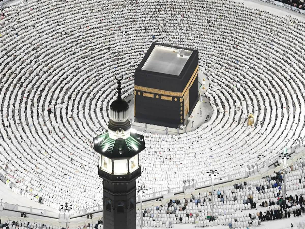 A crowd in white surrounds the Kaaba, a black cubic structure with gold details, in a lit courtyard at night. A green-lit minaret is in the foreground, during a religious gathering, likely Haj or Umrah in Mecca, Saudi Arabia.