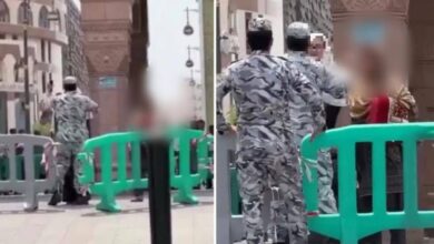Two security officers confront a woman near a barricade outside the Prophet’s Mosque in Madinah, with the mosque and city structures in the background. The image is blurred for privacy.