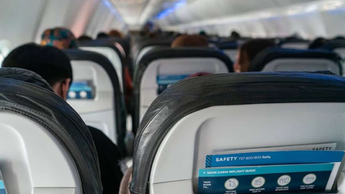 Interior view of an airplane cabin showing passengers seated with safety instruction cards visible in the seat pockets – economy class travel.