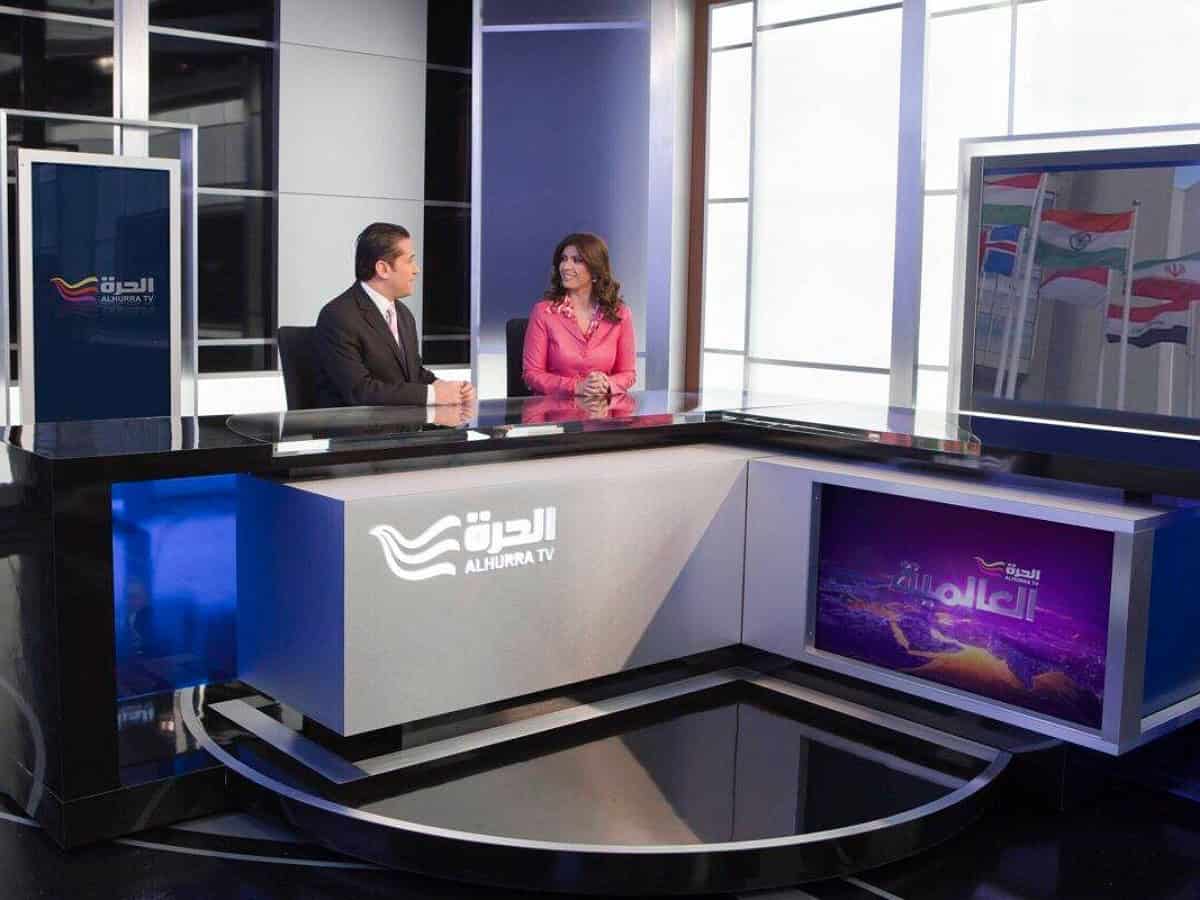Alhurra TV news anchors in a modern studio with a glass desk, digital screens, and international flags in the background.