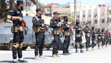 Syrian security forces in black uniforms standing in formation with rifles during a street deployment in a southern suburb of Damascus amid ongoing clashes.