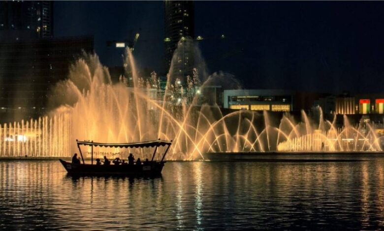 Traditional boat with people sailing on Burj Lake at night with Dubai Fountain water show in the background, lit with golden lights near Dubai Mall, UAE.