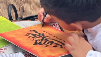 A young child, dressed in a white traditional outfit, is engaged in calligraphy on a vibrant orange and yellow canvas.