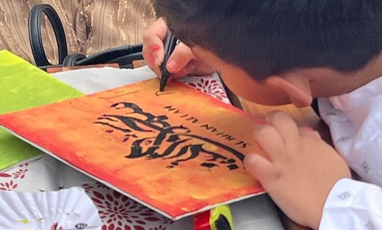 A young child, dressed in a white traditional outfit, is engaged in calligraphy on a vibrant orange and yellow canvas.