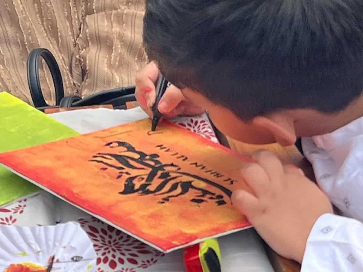A young child, dressed in a white traditional outfit, is engaged in calligraphy on a vibrant orange and yellow canvas.