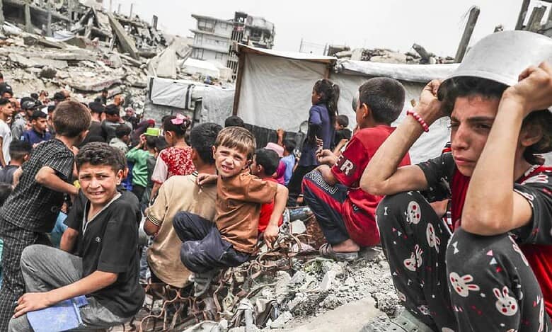 Children sit amid rubble in Gaza Strip, with worried expressions, in a crowded area of debris and destroyed buildings. A child wears a pot as a hat, reflecting distress.