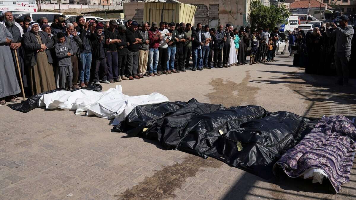 A large group of mourners stands in prayer over several bodies wrapped in black and white body bags, laid out on a paved ground in broad daylight.