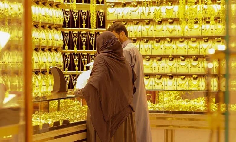 A man and woman in traditional attire shopping for gold jewellery in a well-lit Dubai gold souk. The shop is filled with gold bangles, necklaces, and ornaments displayed in glass counters and wall panels, creating a luxurious, golden ambience.