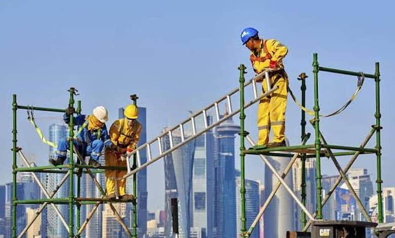 Construction workers on scaffolding at a building site in a Gulf city, wearing safety helmets and high-visibility clothing.